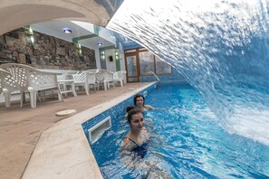Una piscina interior con dos personas nadando, rodeada de sillas de plástico blanco y un muro de piedra.