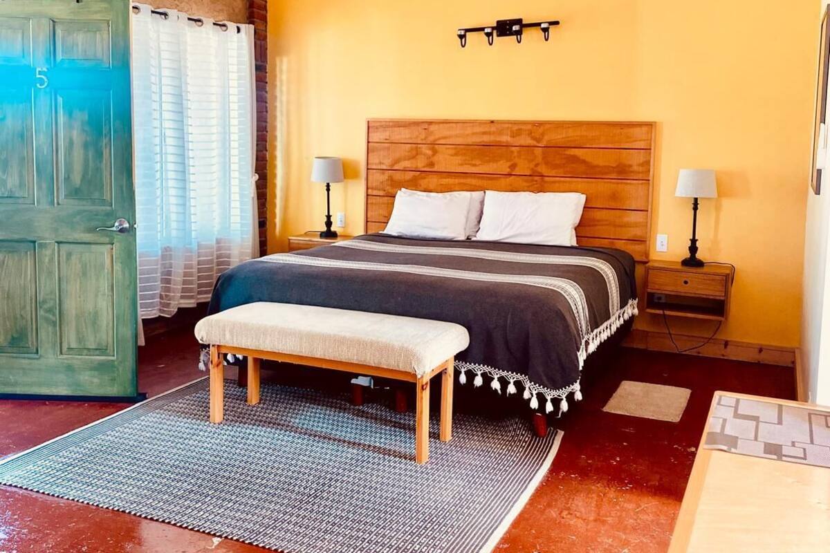 A bedroom with a wooden headboard, a single bed, a bench, two bedside tables with lamps, a ceiling fan, and a door with a number.