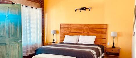Quarto com cabeceira de madeira, uma cama de solteiro, um banquinho, duas mesas de cabeceira com luminárias, um ventilador de teto e uma porta com número.