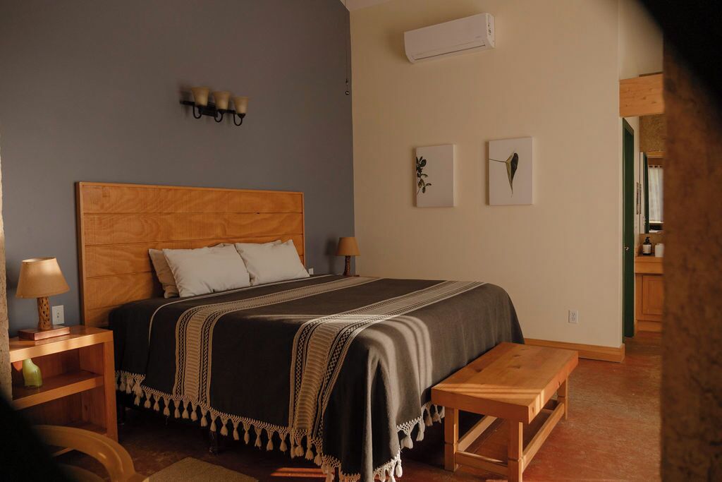 Une chambre à coucher avec une tête de lit en bois, un lit recouvert d’une couverture foncée, des tables de chevet avec des lampes et un banc en bois.
