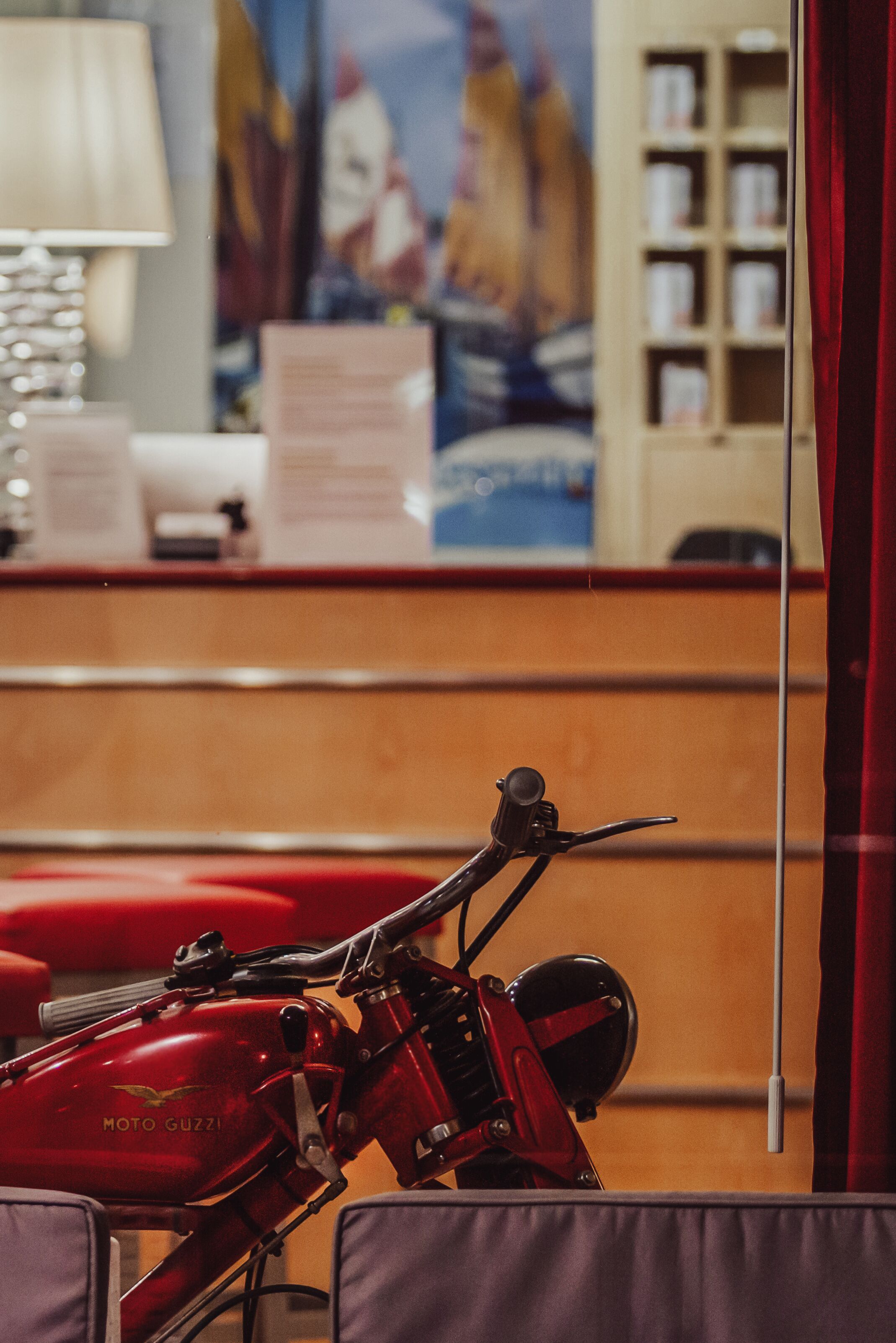A red motorcycle in a room with a wooden reception desk, a lamp, and a poster on the wall.