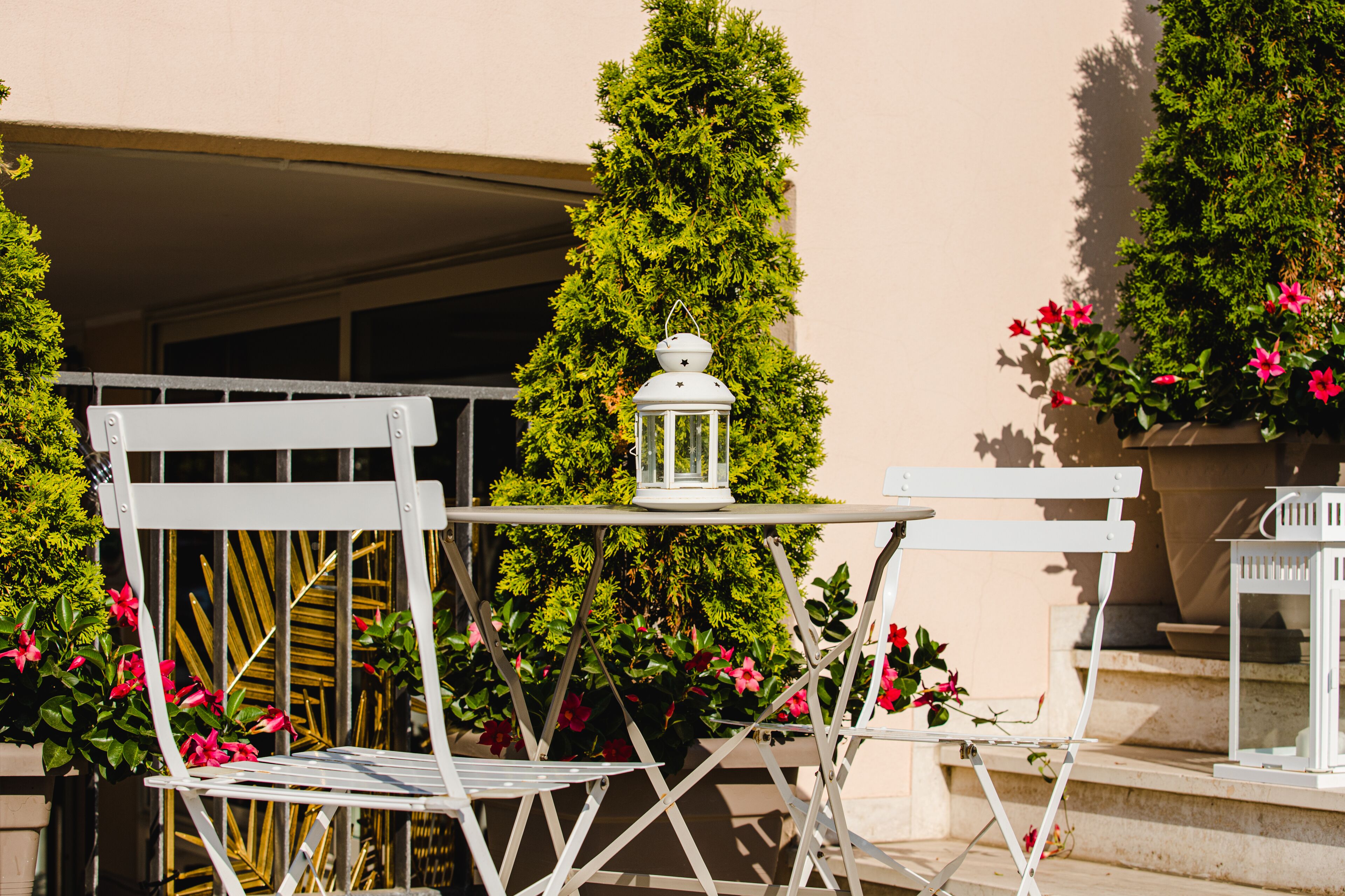 A white metal chair and table set on a balcony with a white lantern, surrounded by green foliage and pink flowers.