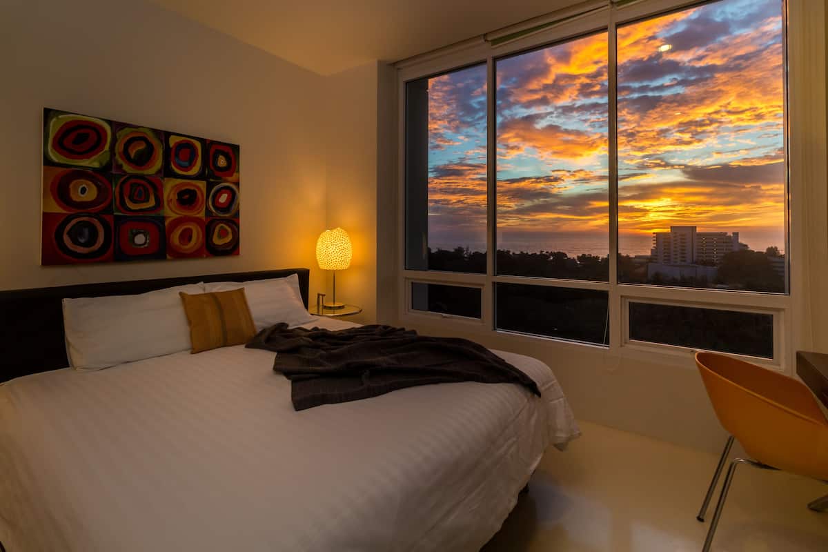 A bedroom with a large bed, a nightstand with a lamp, a chair, and a view of a sunset through a window.