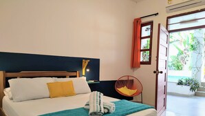 Un lit bien fait, recouvert d’une couverture turquoise et agrémenté d’une serviette enroulée, une tête de lit en bois, un luminaire fixé au mur et une chaise rouge.