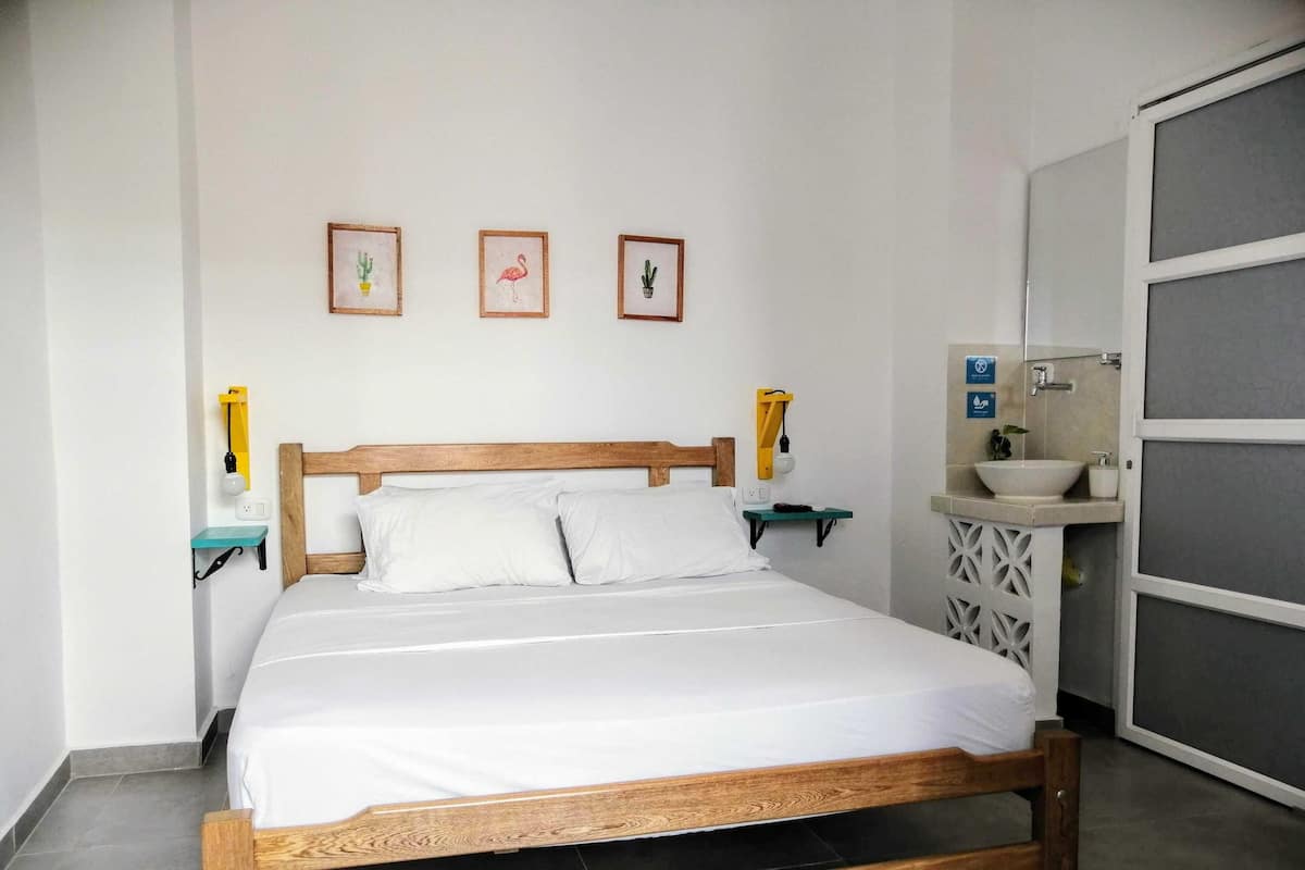 A neatly made bed with a wooden headboard, a bedside table with a lamp, and a small shelf with a plant. There are three framed pictures on the wall above the bed.