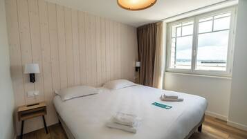A hotel room with a wooden panel wall, a bed with white linens, a nightstand with a lamp, and a window with a view of the sea.