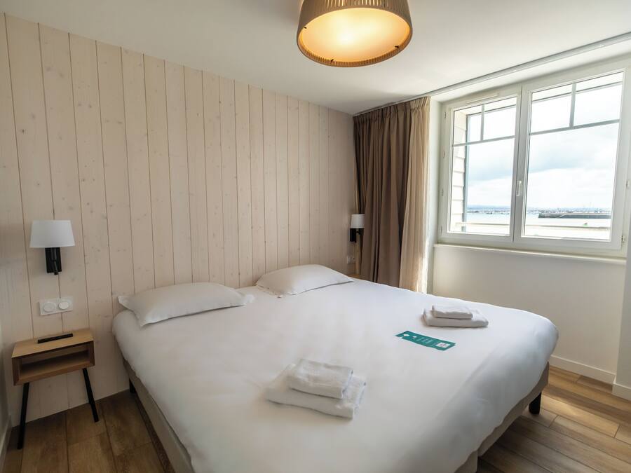 A hotel room with a wooden panel wall, a bed with white linens, a nightstand with a lamp, and a window with a view of the sea.