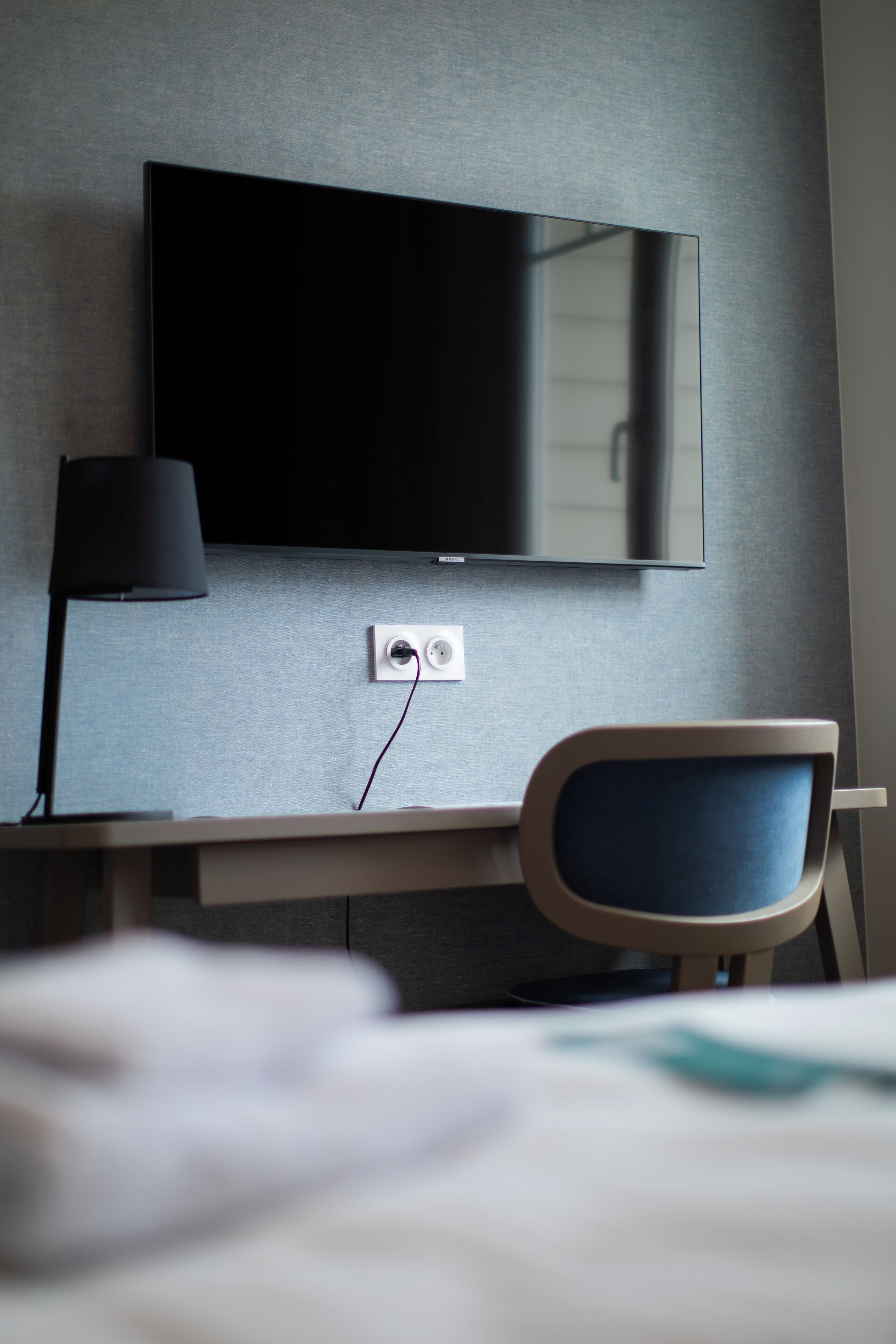 A modern hotel room with a flat-screen TV mounted on the wall, a desk with a lamp, and a chair.