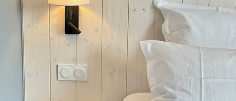 A bedroom with a wooden nightstand, a wall-mounted lamp, and a bed with white bedding.