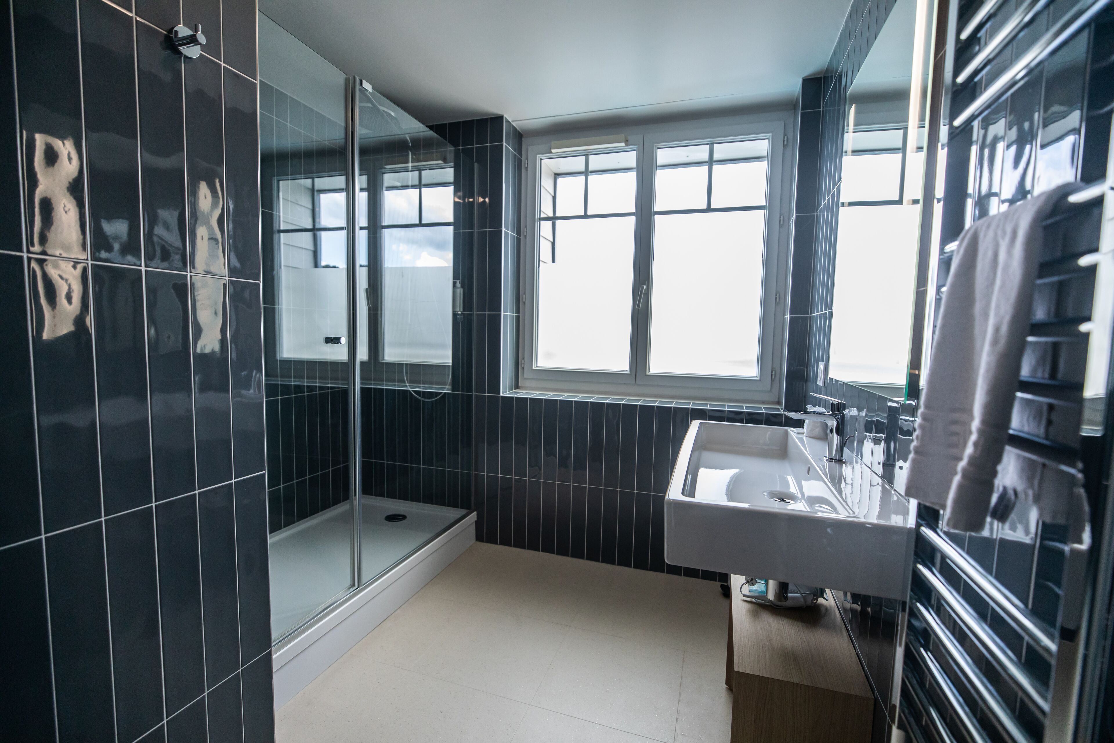 A modern bathroom with dark tiled walls, a glass shower enclosure, a white sink, and a window providing natural light.