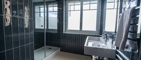 Un baño moderno con paredes de azulejos oscuros, cabina de ducha con mampara de vidrio, lavamanos blanco y ventana que permite la entrada de luz natural.
