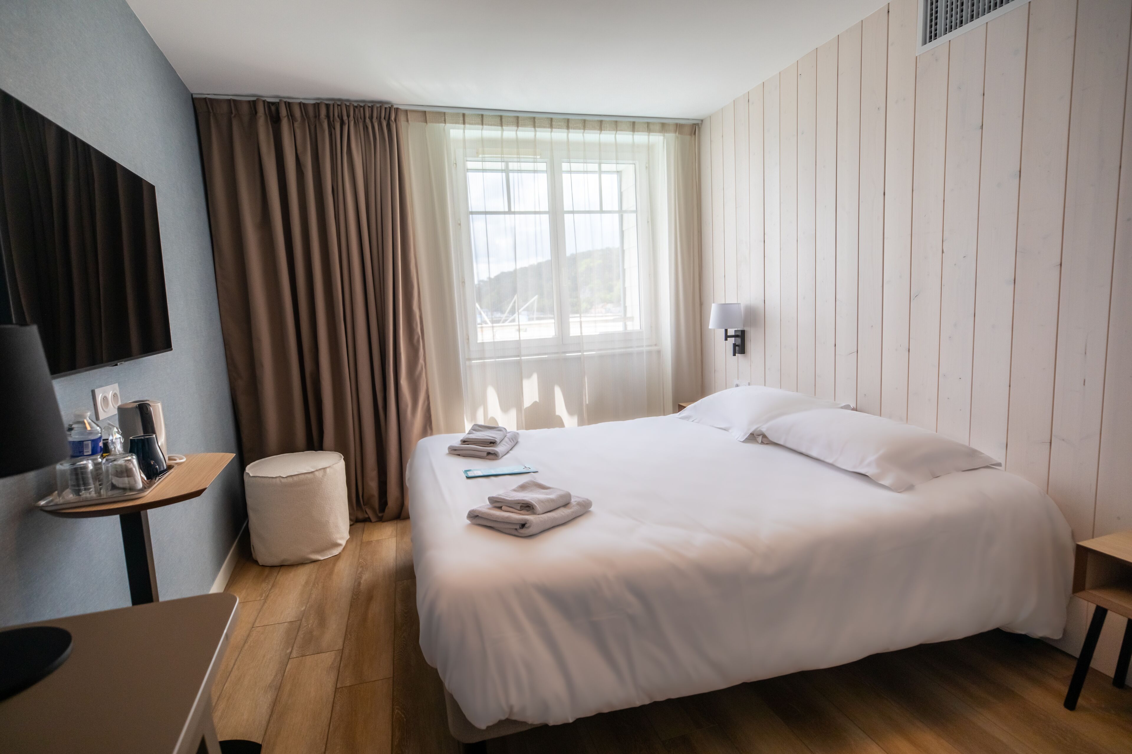 Une chambre d’hôtel avec un grand lit, un bureau avec une lampe, une petite table sur laquelle se trouve un plateau avec divers objets, et une fenêtre avec des rideaux.