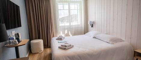 Une chambre d’hôtel avec un grand lit, un bureau avec une lampe, une petite table sur laquelle se trouve un plateau avec divers objets, et une fenêtre avec des rideaux.