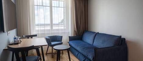 A modern living room with a blue sofa, a wooden dining table with chairs, and a large window with curtains.