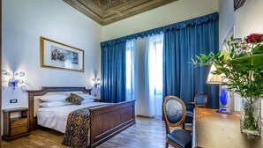 A hotel room with a wooden bed, a bedside table, a chair, a desk, a lamp, a vase with flowers, and a framed picture on the wall.