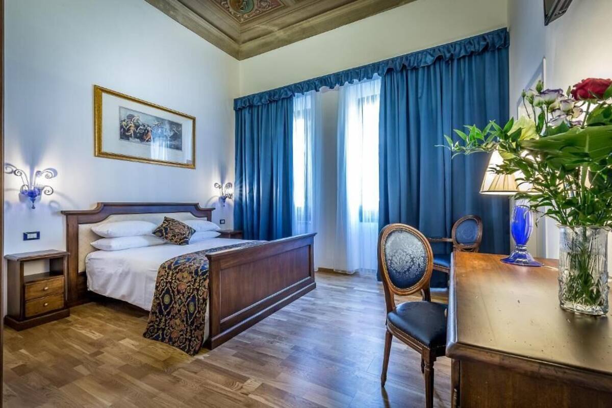 A hotel room with a wooden bed, a bedside table, a chair, a desk, a lamp, a vase with flowers, and a framed picture on the wall.