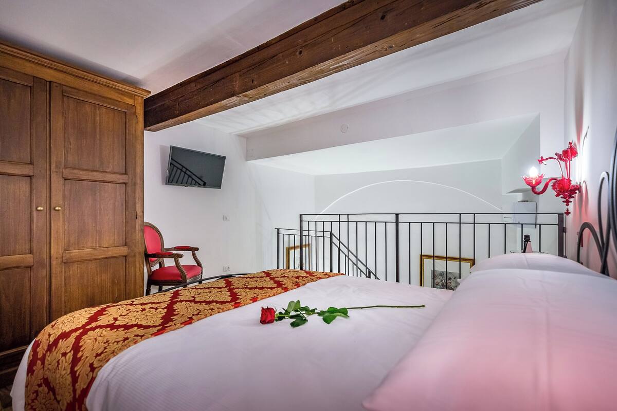 A bedroom with a wooden wardrobe, a red chair, a bed with a white and red bedspread, a single rose, and a staircase.