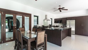 A modern kitchen with dark cabinetry, a central island, and a dining area with a glass table and chairs. - Villa Frangipani by Lofty (Rawai)