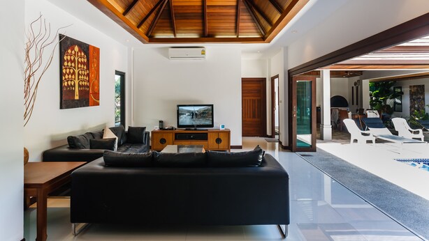 A modern living room with a black sofa, wooden entertainment unit, and a television. There is a pool area visible through an open doorway. - Villa Frangipani by Lofty (Rawai)