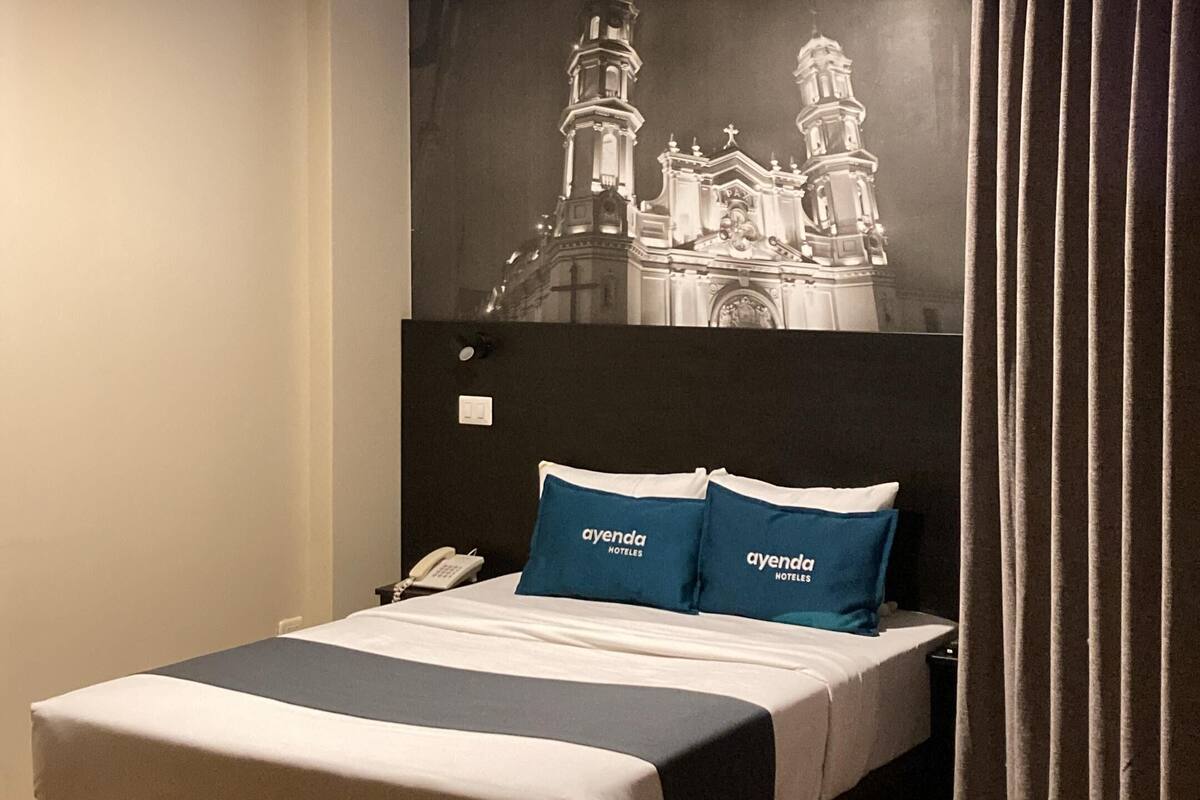 Habitación de hotel con una cama grande, cabecera con un mural de un paisaje urbano, un teléfono y una cortina.