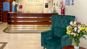 Zona de recepción de un hotel con un mostrador de madera, un sillón verde y un jarrón con flores.