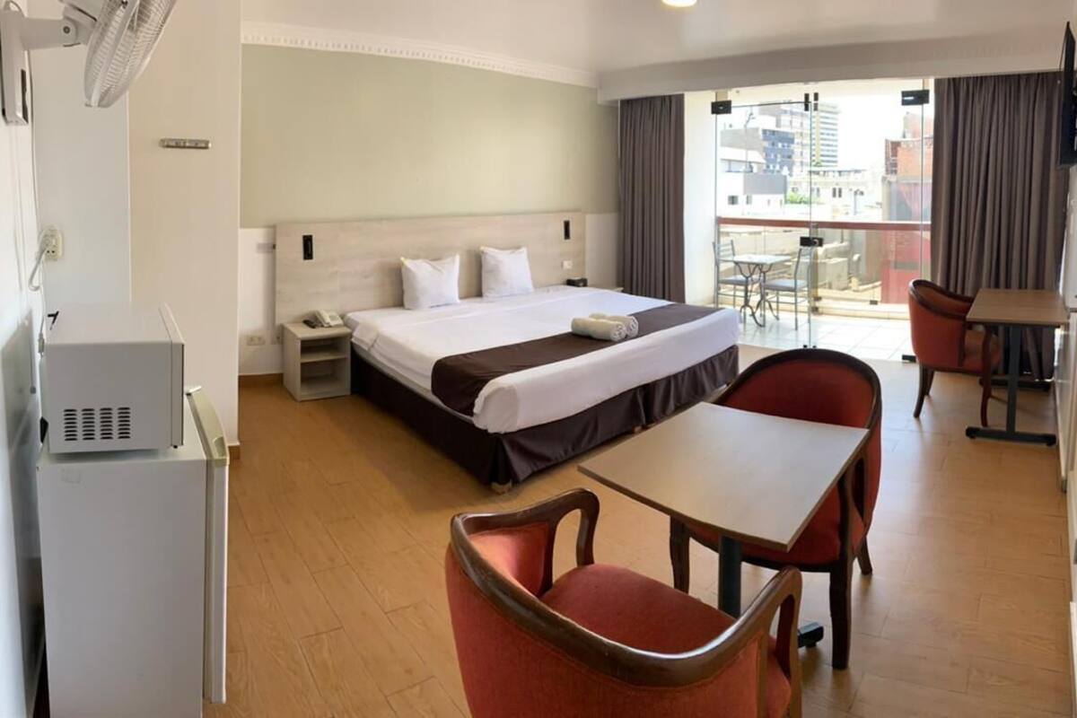 Habitación de hotel con una cama, un escritorio y una silla. Hay una ventana con cortinas, un ventilador y un televisor en la pared.
