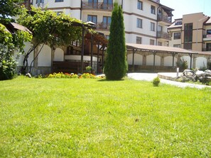 A courtyard with a well-maintained lawn, a tall coniferous tree, and a building with multiple balconies and windows. - Olymp Hotel (Bansko)