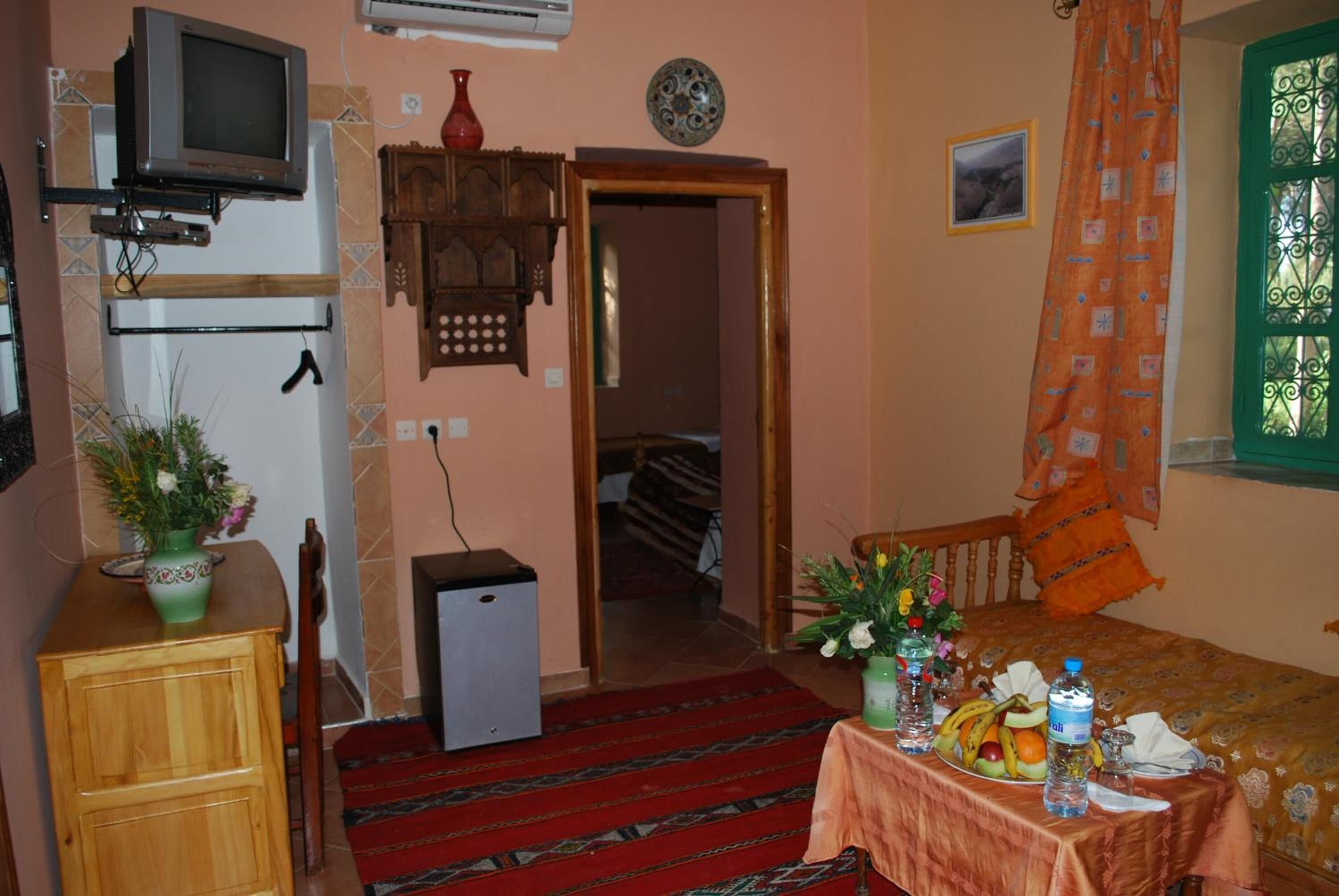 A small, cozy room with a bed, a table with food and drinks, a TV, a small fridge, and a door leading to another room.