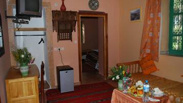 A small, cozy room with a bed, a table with food and drinks, a TV, a small fridge, and a door leading to another room.