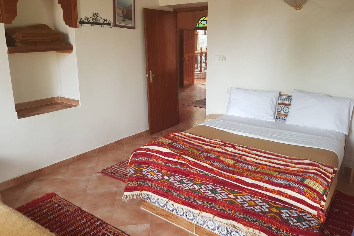 Um quarto com uma cama, um cobertor estampado, uma prateleira de madeira, uma porta e um aplique na parede.