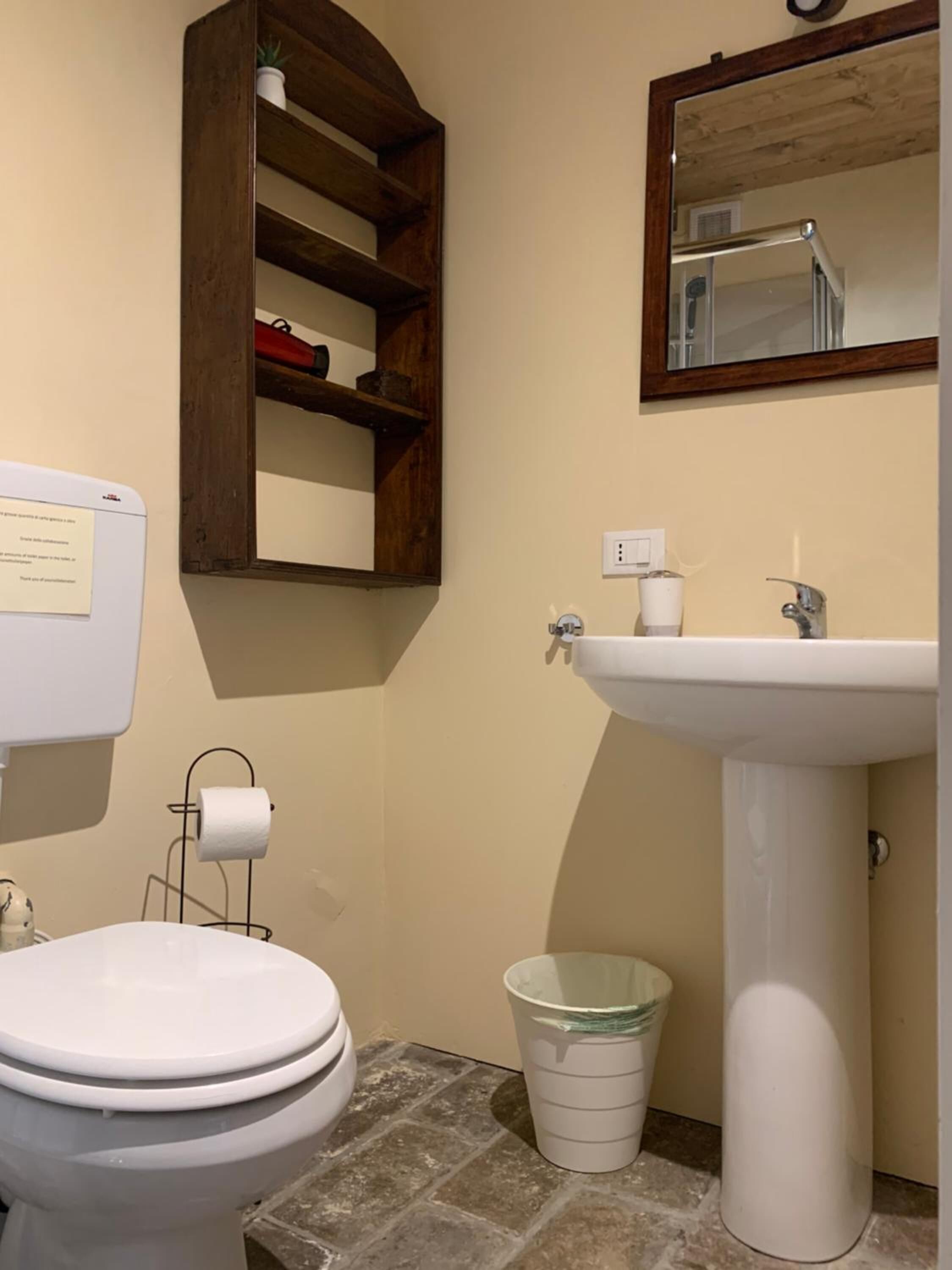 A bathroom with a toilet, a sink, a mirror, a wooden shelf, and a trash bin.