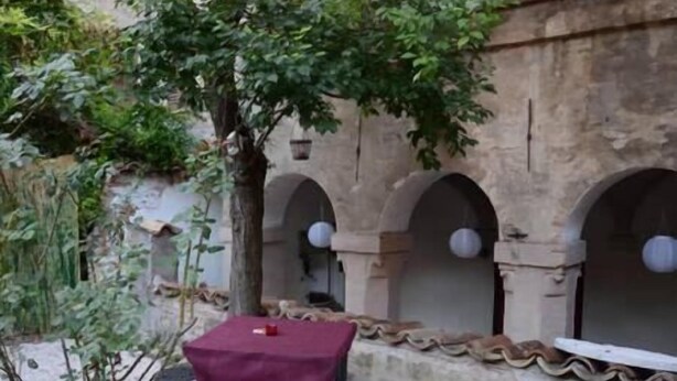 Uma área externa com mesas e cadeiras, uma árvore e um edifício com arcos e lanternas.