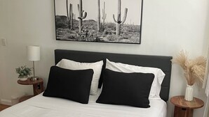 Una cama bien hecha con ropa de cama blanco y negro, una obra de arte con temática de cactus en la pared, una lámpara de noche y una planta pequeña sobre una mesita de noche.