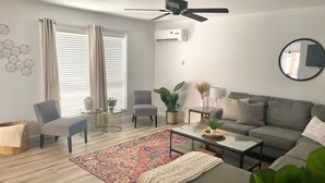 Una sala de estar moderna con un sofá gris, dos sillones, una mesa de centro y un tapete rojo estampado. Hay un ventilador de techo, un equipo de aire acondicionado y un espejo redondo en la pared.