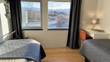 Una habitación con dos camas, un escritorio y una silla. Hay dos ventanas con vistas a árboles y montañas.