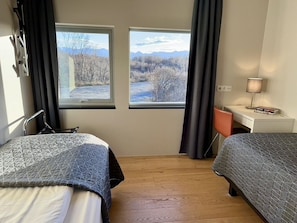 Quarto com duas camas, uma escrivaninha e uma cadeira. Duas janelas oferecem vista para árvores e montanhas.