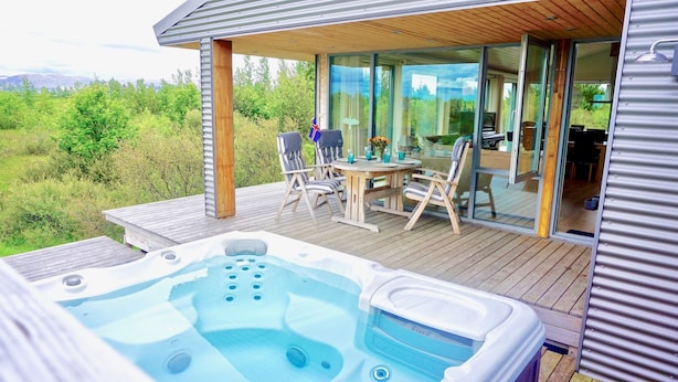 A wooden deck with a hot tub, outdoor seating, and a glass-enclosed dining area with a view of the surrounding landscape. - Luxury House with a Cozy Guesthouse + Hot Tub close to Snæfellsnes National Park (Borgarnes)