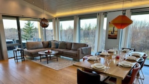 A modern dining area with a wooden table set for six, a sofa, and a view of the outdoors. - Luxury House with a Cozy Guesthouse + Hot Tub close to Snæfellsnes National Park (Borgarnes)