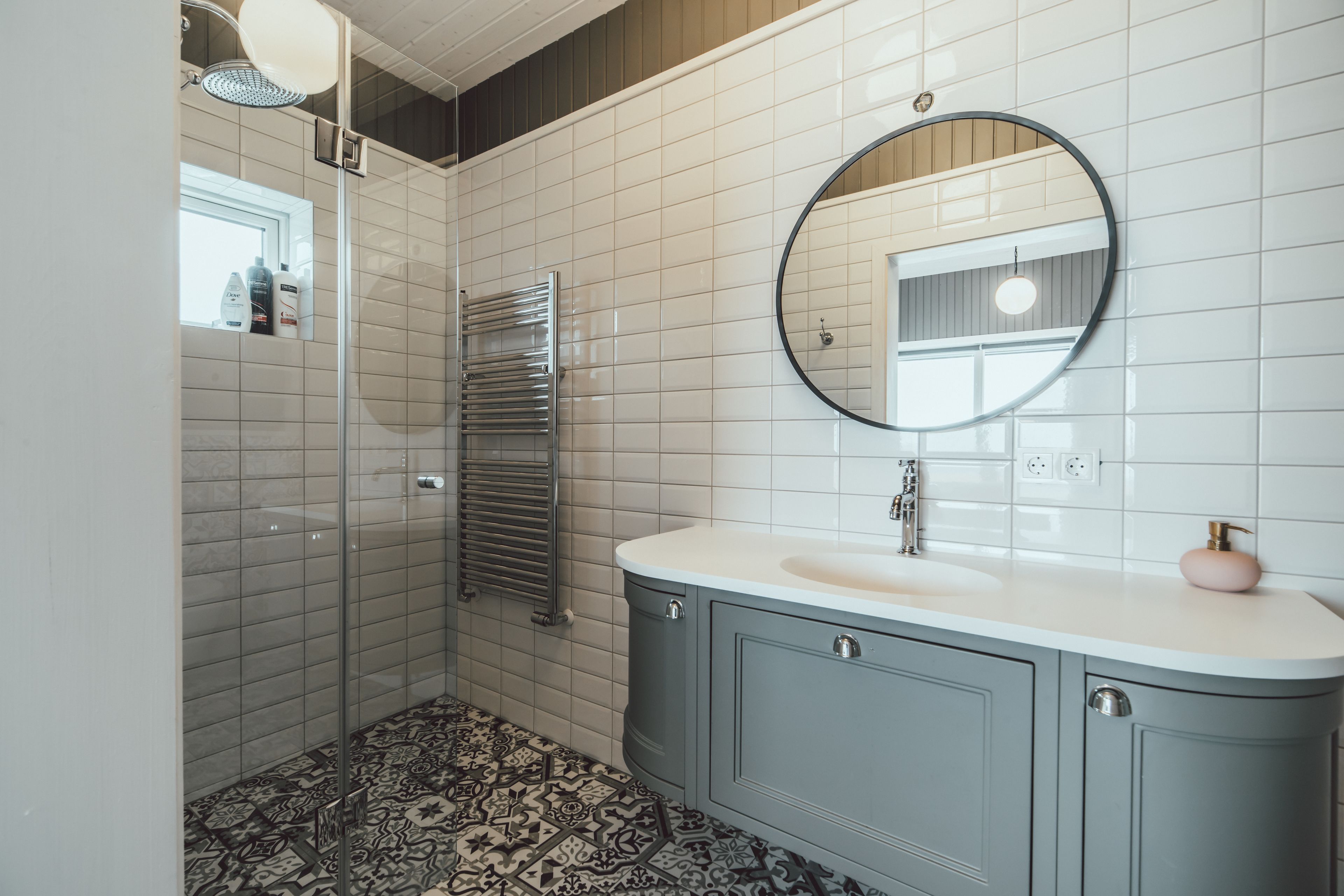 A bathroom with a white tiled wall, a round mirror, a sink with a pink soap dispenser, and a patterned floor.