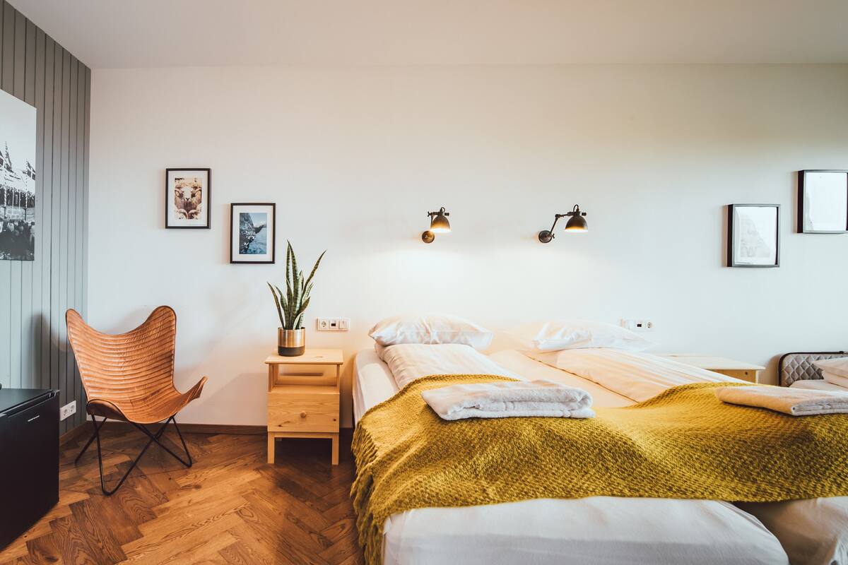 A modern hotel room with a wooden floor, a bed with a yellow blanket, a chair, a nightstand, and wall-mounted lights.