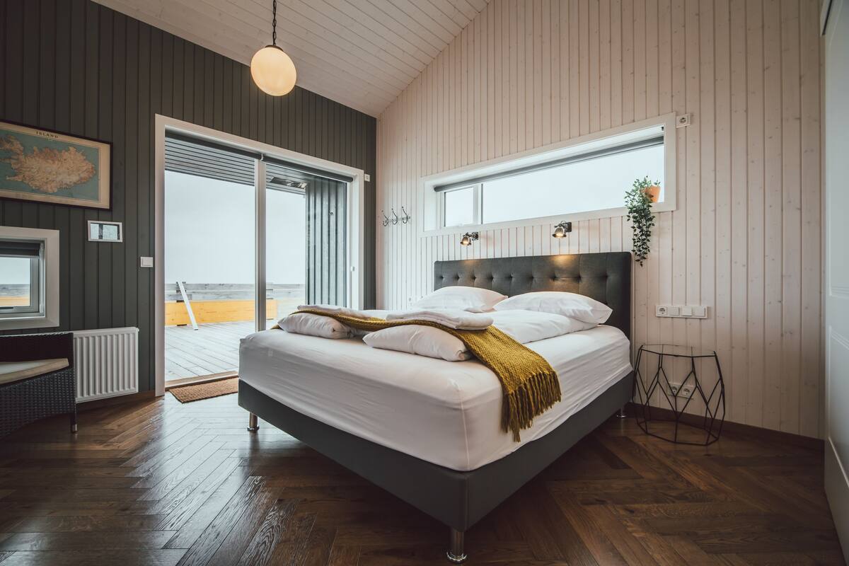 A bedroom with a large bed, wooden flooring, a view through a sliding door, and a pendant light.