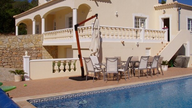 A poolside area with a white umbrella, lounge chairs, and a stone wall. - Beautiful villa, with private pool, set in hills with spectacular views  (Loule)