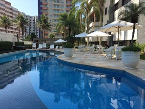 A swimming pool with lounge chairs and umbrellas, surrounded by palm trees and modern buildings. - Riviera de São Lourenço - Module VI, 250m from the beach. (Bertioga)
