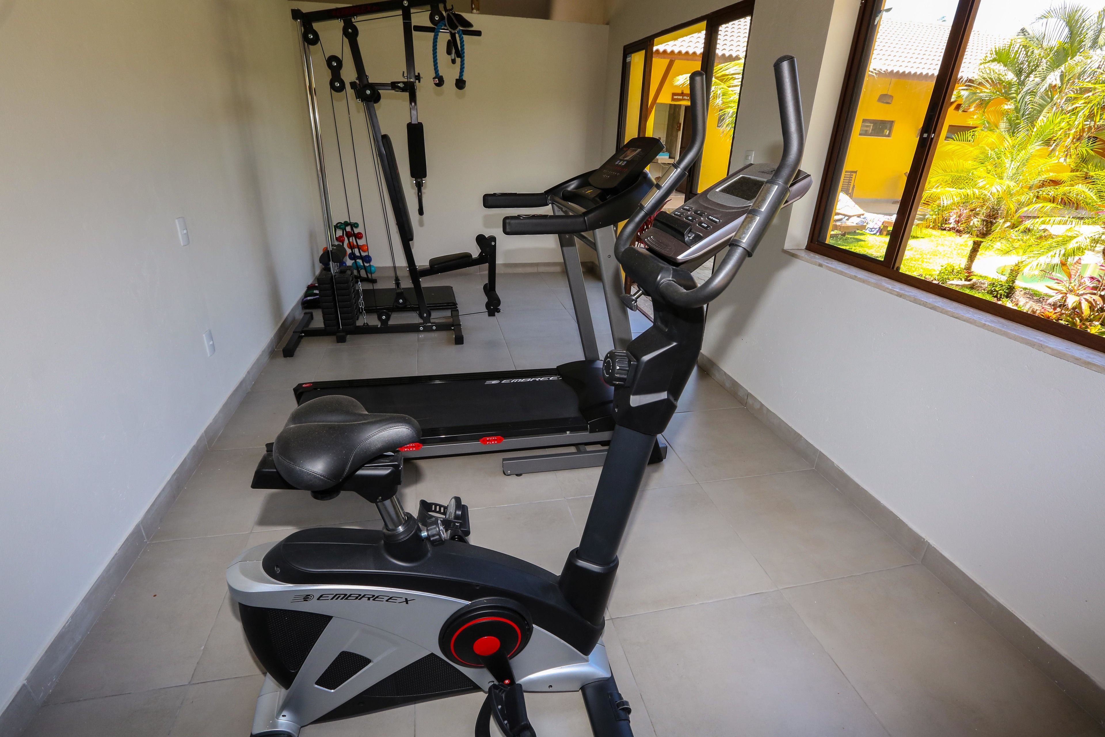 A gym with cardio machines, weightlifting equipment, and a window with a view of a yellow building and greenery.