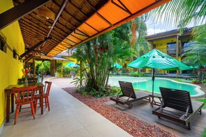 A poolside area with wooden chairs and tables, surrounded by tropical plants and a pool.