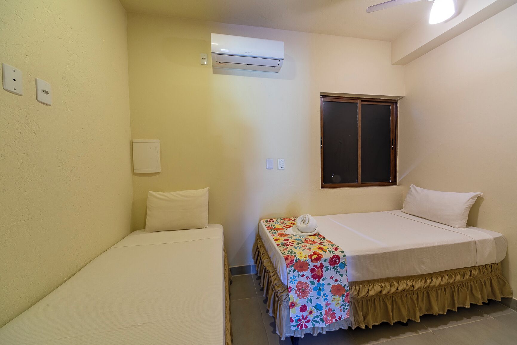 Quarto de hotel pequeno e individual, com uma cama, janela, unidade de ar condicionado e luminária fixada na parede.