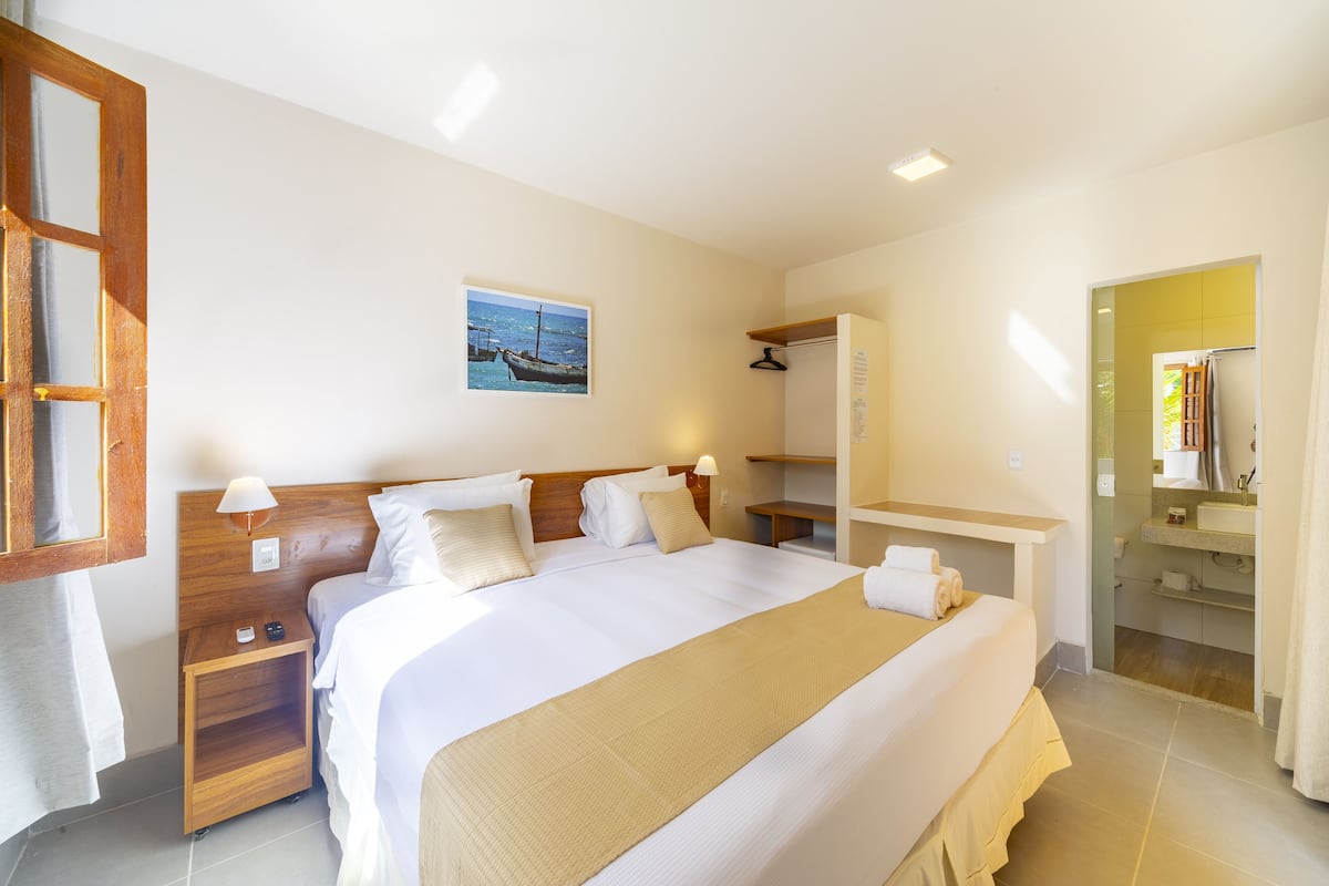 Ein Hotelzimmer mit einem großen Bett, einem Holzkopfstück, Nachttischen, einem Beistelltisch, einer Wandlampe, einem Handtuch, einem Bild von einem Boot und einem durchgegangene Tür sichtbaren Badezimmer.