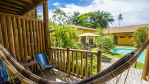 A hammock on a wooden deck with a view of a swimming pool and a yellow building. - Pousada Tropicarim (Camaçari)
