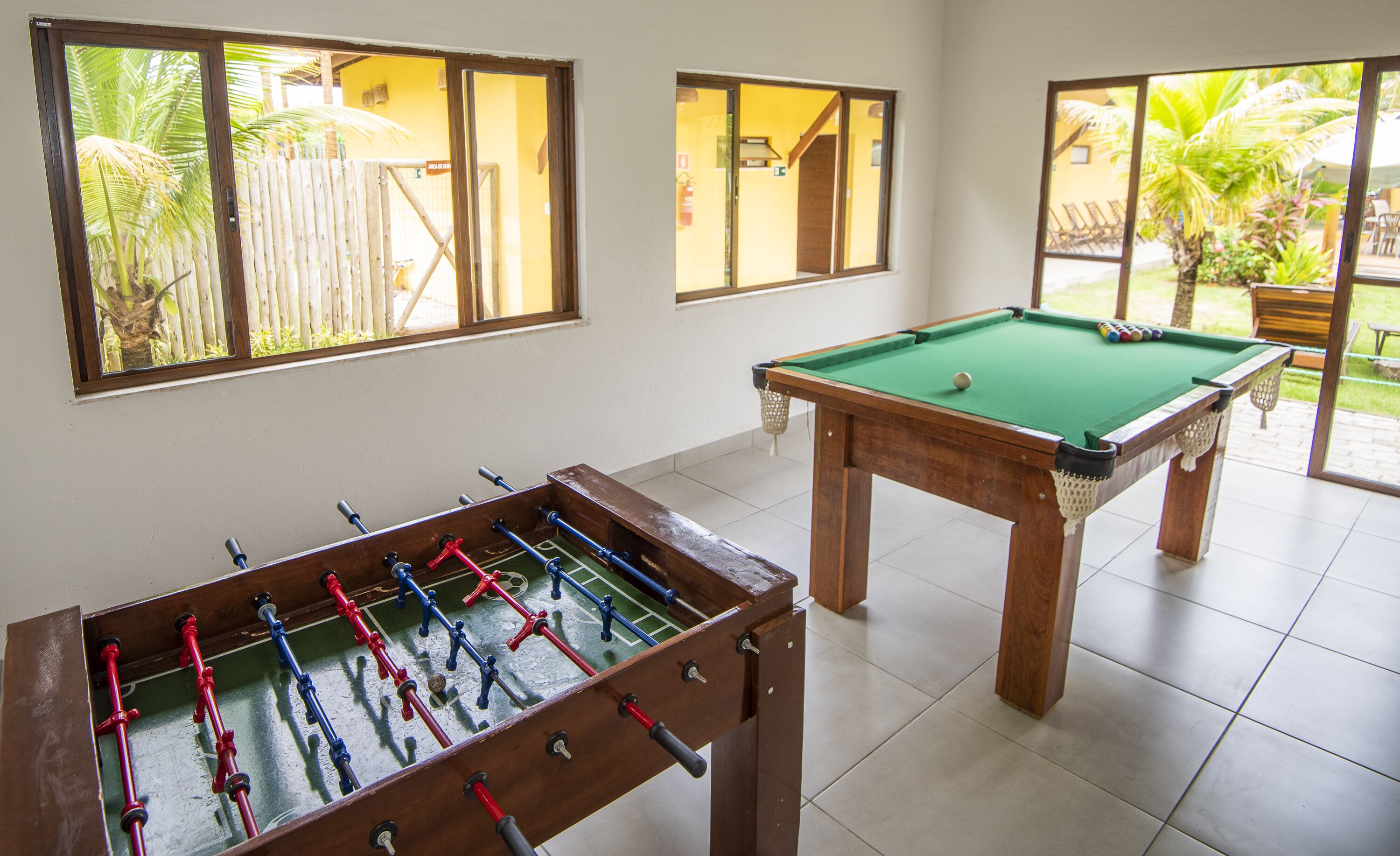 A room with a foosball table, a pool table, and large windows with views of palm trees.