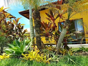 A yellow building with a garden in front, including various tropical plants and a palm tree. - Pousada Tropicarim (Camaçari)